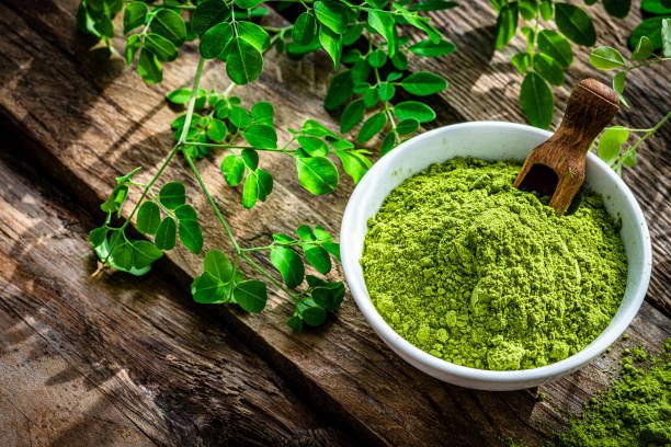 Nutritional supplement: high angle view of Moringa oleifera powder in a white bowl shot on rustic wooden table. Green Moringa leaves are around the bowl. High resolution 42Mp studio digital capture taken with Sony A7rII and Sony FE 90mm f2.8 macro G OSS lens
