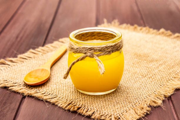 Pure or desi ghee (ghi), clarified melted butter. Healthy fats bulletproof diet concept or paleo style plan. Glass jar, wooden spoon on vintage sackcloth. Wooden boards background, copy space close up